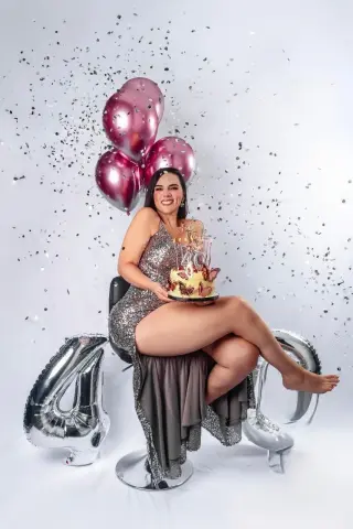 Smiling woman in a sparkly silver dress holding a birthday cake with pink balloons and silver number 40 decor.