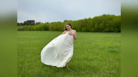 Smiling woman in a flowing white dress walking through a lush green meadow under a soft sky.