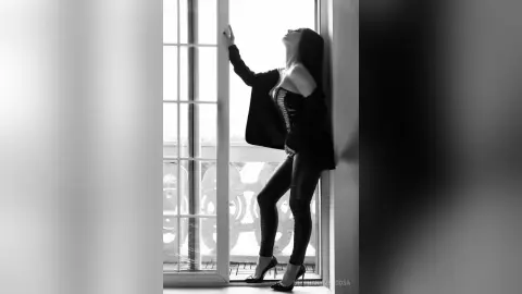 Moody black and white portrait of a woman in a black bodysuit leaning against a bright window frame.