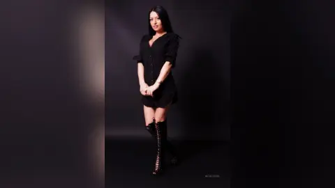 NicolleSun poses in a black button-down shirt and black lace-up thigh-high boots against a dark background.