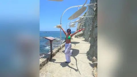 Cheerful woman in a green bikini top and white linen pants enjoying a sunny day on a coastal stone walkway.