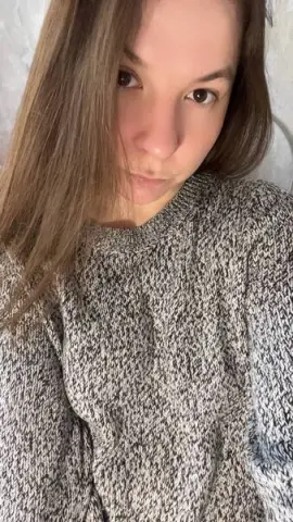 Young woman with long brown hair wearing a cozy grey knit sweater in a bright indoor setting.