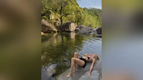 Blonde woman in a black bikini relaxing on rocks by a serene forest river in nature.