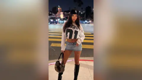 SamanthaBrazer wears a white long-sleeve crop top and denim mini skirt while standing on a city street at night, holding a patterned handbag.