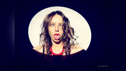 Playful woman with curly hair and red lipstick posing in a circular spotlight against a dark background.