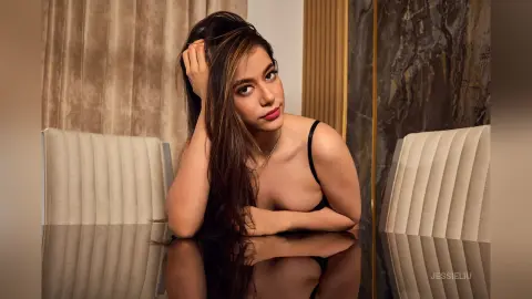 JessieLiu leans forward over a reflective table, wearing a black bra and a silver necklace while running her hand through her long brown hair.