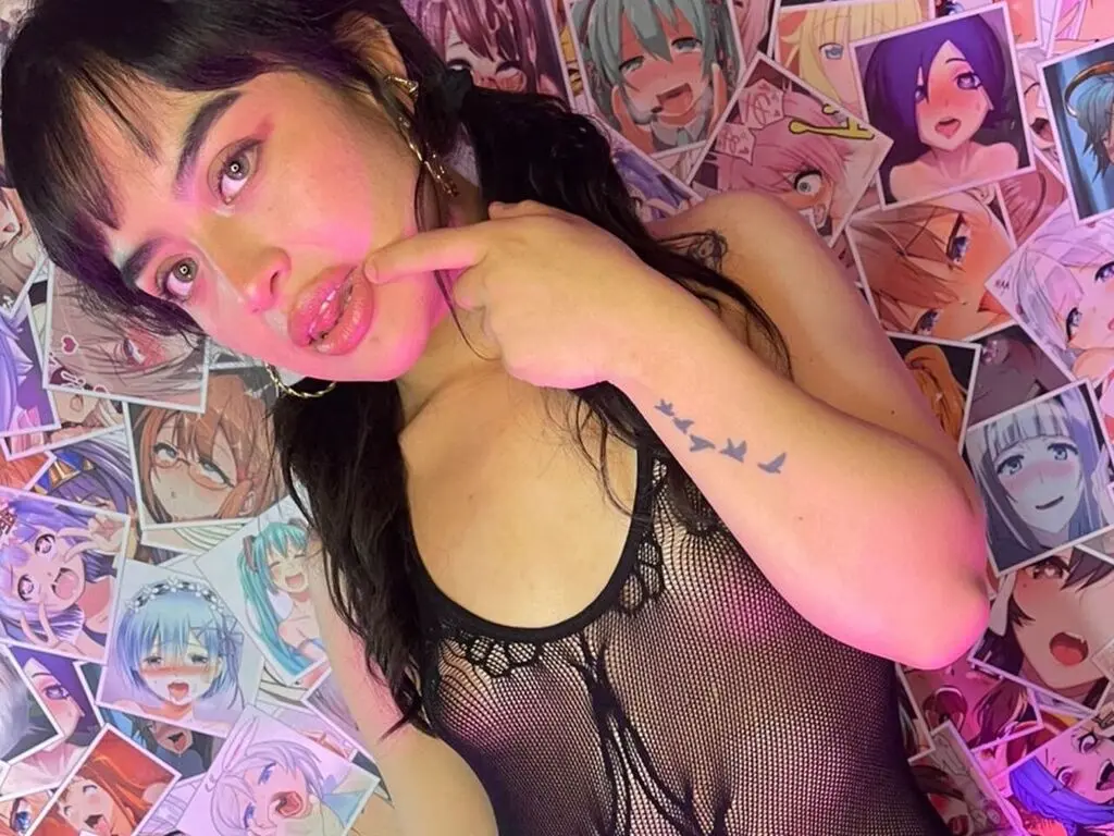 Young woman in a black mesh top posing playfully against a wall of colorful anime posters.