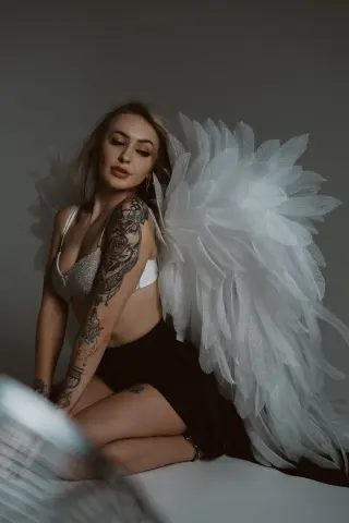 Tattooed woman in a white bikini and large feathery angel wings posing against a grey studio background.