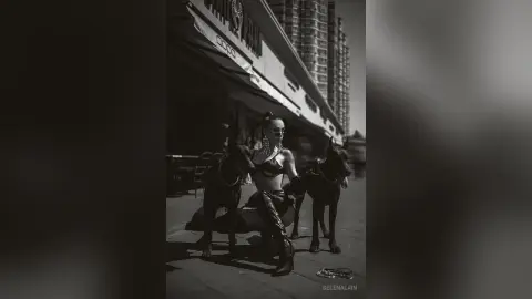 Stylish woman in a black bikini and leather boots posing with two dogs on a sunny city street.