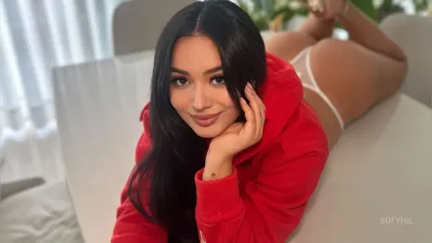 Smiling brunette woman in a red hoodie relaxing on a white sofa in a bright, cozy indoor setting.