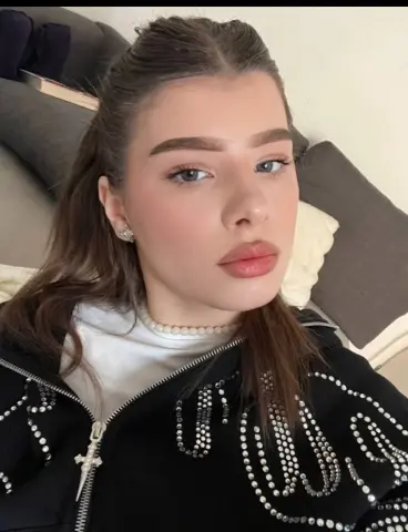 Young woman with long brown hair wearing a white top and black studded jacket in a cozy indoor setting.