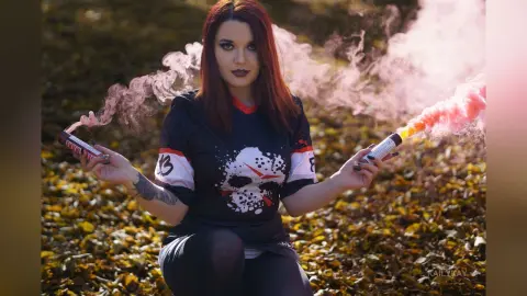 Red-haired woman in a black graphic t-shirt holding pink smoke bombs in an autumn forest setting.