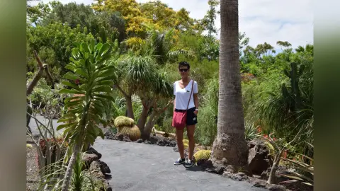 Woman in white t-shirt and red shorts standing on a garden path surrounded by lush tropical plants and palm trees.