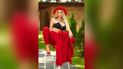Anastasia poses outdoors in a black lace bra and red trousers, leaning forward over a white stool with her long blonde hair down.