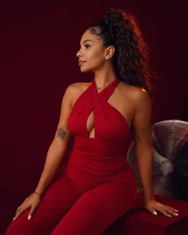 ScarlettPearls — ScarlettPearls poses in a red halter jumpsuit against a dark red background.