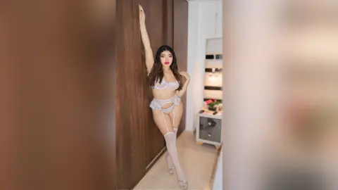 Glamorous woman in white lace lingerie and stockings posing in a modern, brightly lit room.