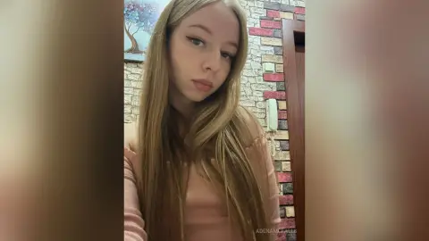 AdenaMcfalls poses with her long blonde hair down, wearing a light pink long-sleeved top against a brick wall background.