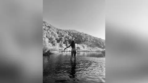 Black and white shot of a woman standing in a calm lake with mountains in the background, peaceful mood.