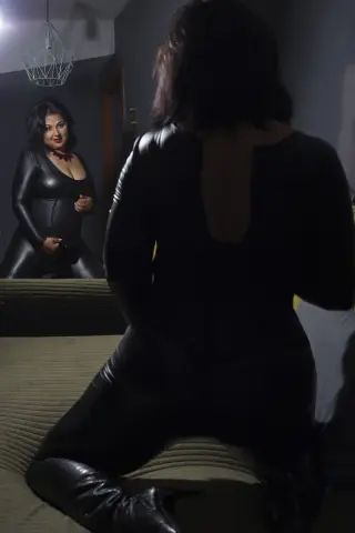 Curvy woman in a sleek black bodysuit sitting in a dimly lit room, viewed through a mirror reflection.