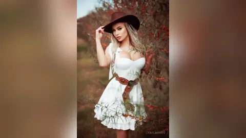 Blonde woman in a white sundress and black cowboy hat posing in a rustic garden setting.