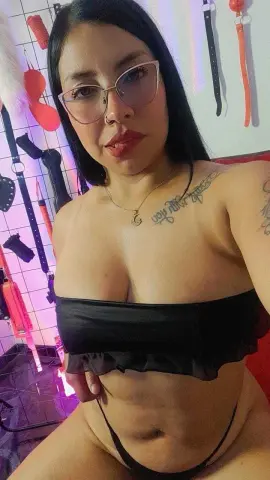 Woman in black bikini top with tattoos posing in a room with colorful neon lighting.