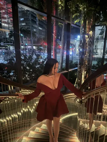 Elegant woman in a burgundy cocktail dress posing on a modern glass staircase at night.