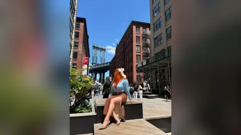 Redheaded woman in a blue top and brown skirt sitting on a city terrace with Manhattan Bridge in the background.