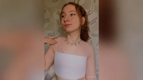 MadgeRemerez poses in a white tank top and heart necklace, showing her light brown hair and shoulder tattoo against a patterned wall.