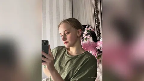 Young woman in an olive green t-shirt taking a mirror selfie in a cozy bedroom with pink floral decor.
