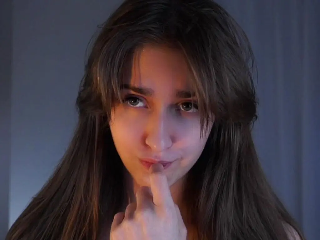 Young woman with long brown hair looking thoughtfully at the camera in soft, moody indoor lighting.