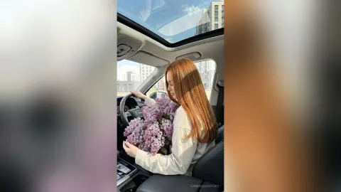 Redheaded woman in a white shirt holding a large bouquet of purple lilacs inside a car with a sunroof.