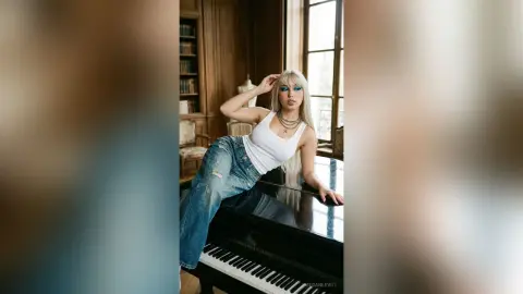 Blonde woman in a white tank top and denim jeans posing elegantly by a grand piano in a classic library.