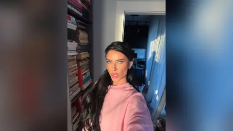 Woman with long dark hair in a pink top posing in a cozy room with a bookshelf background.