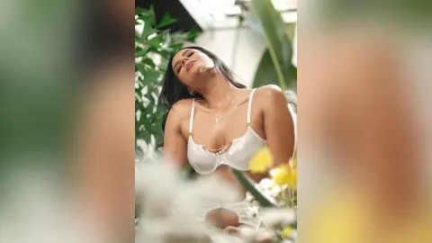 Serene woman in white lace lingerie relaxing amidst lush green tropical plants in a bright, dreamy setting.
