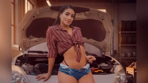 Smiling woman in a red striped tie-front shirt and denim shorts leaning against a car with an open hood.