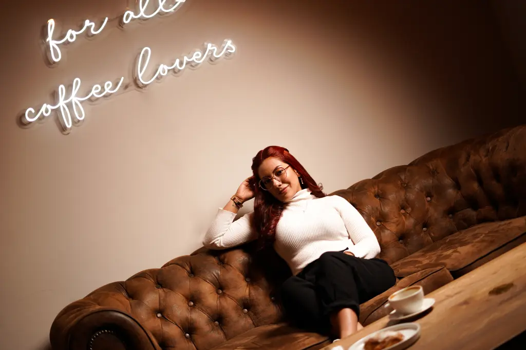 desk_desire — Smiling woman in a white sweater relaxing on a brown leather sofa in a cozy cafe with neon coffee sign.