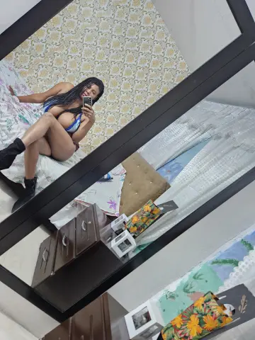 Smiling woman in blue bikini sitting on a bed in a bright bedroom taking a mirror selfie.