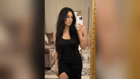 Young woman in a sleek black mini dress taking a mirror selfie in a modern living room.