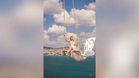 Anita sits on a wooden swing wearing a white dress against a blue sky and ocean background.
