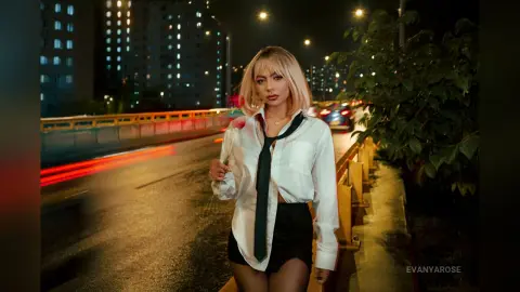 Blonde woman in a white shirt and black tie standing on a city street at night with motion blur lights.