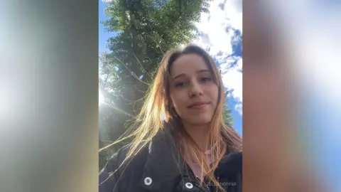 Young woman in a black puffer jacket smiling outdoors under bright sunlight and green trees.