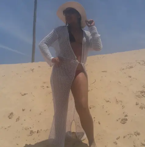 Woman in white crochet beach cover-up and sun hat standing on a sunny sand dune.