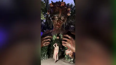 Woman in a beige bodysuit standing before a massive, mystical wooden tree sculpture in a lush jungle setting.