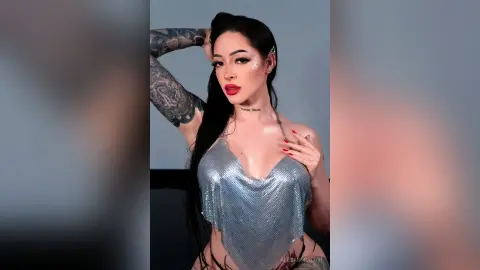 Tattooed woman in a shimmering silver bodysuit posing confidently against a minimalist grey studio background.