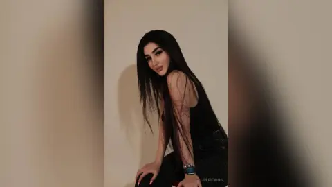 JuliDeming with long dark hair poses in a black tank top and black jeans against a plain wall.