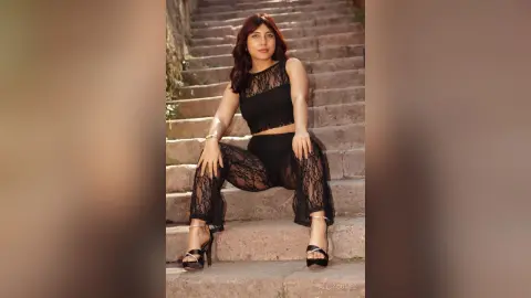 Stylish woman in a black lace crop top and matching lace trousers sitting on stone stairs in a moody outdoor setting.