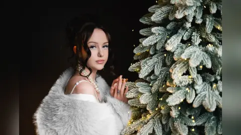 Elegant woman in a white dress and faux fur coat posing next to a glowing frosted Christmas tree.