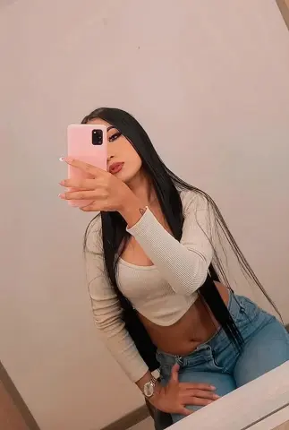 Young woman with long dark hair in a white ribbed crop top and blue jeans taking a mirror selfie.