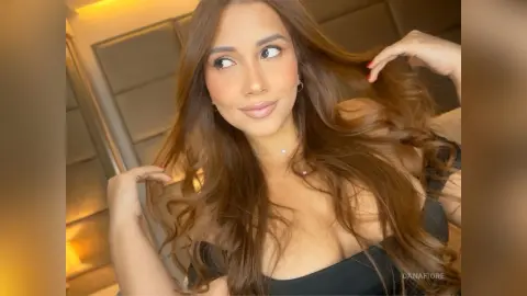 Beautiful woman with long wavy brown hair wearing a black top in a warm, indoor setting.