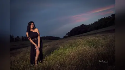 MissyJolie stands in a grassy field wearing a long black dress with a cut-out detail at her waist.